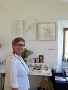 Eine Person steht in einem Ausstellungsraum vor einer Wand mit gerahmten Kunstwerken. Auf einem Tisch vor ihr liegen weitere kleine Kunstobjekte und Karten. Die Person trägt eine Brille und rote Ohrringe. Im Hintergrund ist ein Fenster.