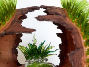 Ein Holzstück mit einem runden Hohlraum steht vertikal, durch dessen Öffnung grünes, grasähnliches Glas sichtbar ist. Oben wachsen ebenfalls grüne Glasstrukturen, die Gras ähneln.