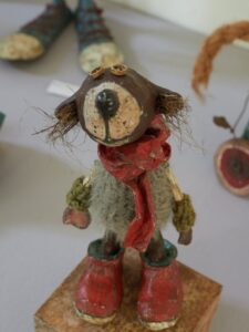 Eine handgefertigte Figur aus verschiedenen Materialien steht aufrecht. Sie hat große rote Stiefel, einen roten Schal und ein tierähnliches Gesicht mit Knopfaugen und borstig wirkendem Fell. Im Hintergrund sind verschwommen weitere Objekte zu sehen.
