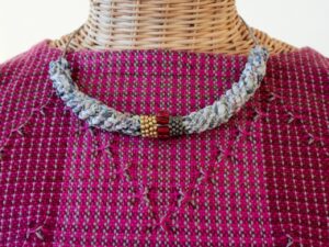 Ein Halskettendetail auf einem Webständer. Die Kette besteht aus geflochtenen blauen Stoffstreifen mit einem Abschnitt aus braunen und goldenen Perlen. Der Hintergrund zeigt ein pink-lila kariertes Muster.